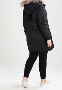 Černá dlouhá parka s kapucí z umělé kožešiny, elastickým pasem a dvěma bočními kapsami, v kombinaci s černými legínami a světlými teniskami.