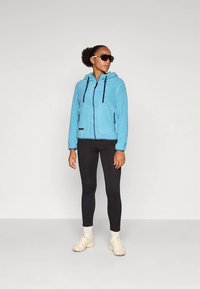 Modra flisasta jopa z zadrgo z rahlo teksturirano površino, z dvema sprednjima žepoma, črnimi vrvicami, v kombinaciji s črnimi legicami in bež supergami.