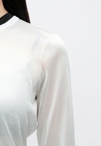 Balts viegls garām piedurknēm tops ar fakturētu audumu un melnu ribotu apkakli. Materiāls šķiet caurspīdīgs ar smalku spīdumu.