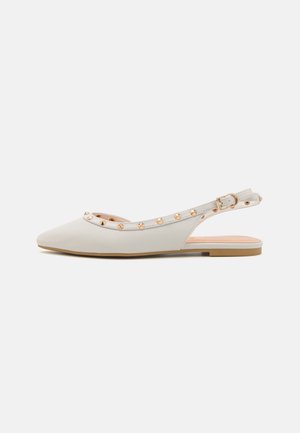 Hvid flad slingback sko med spids tå og gyldne nitteudsmykninger langs kanten og ankelstroppen.