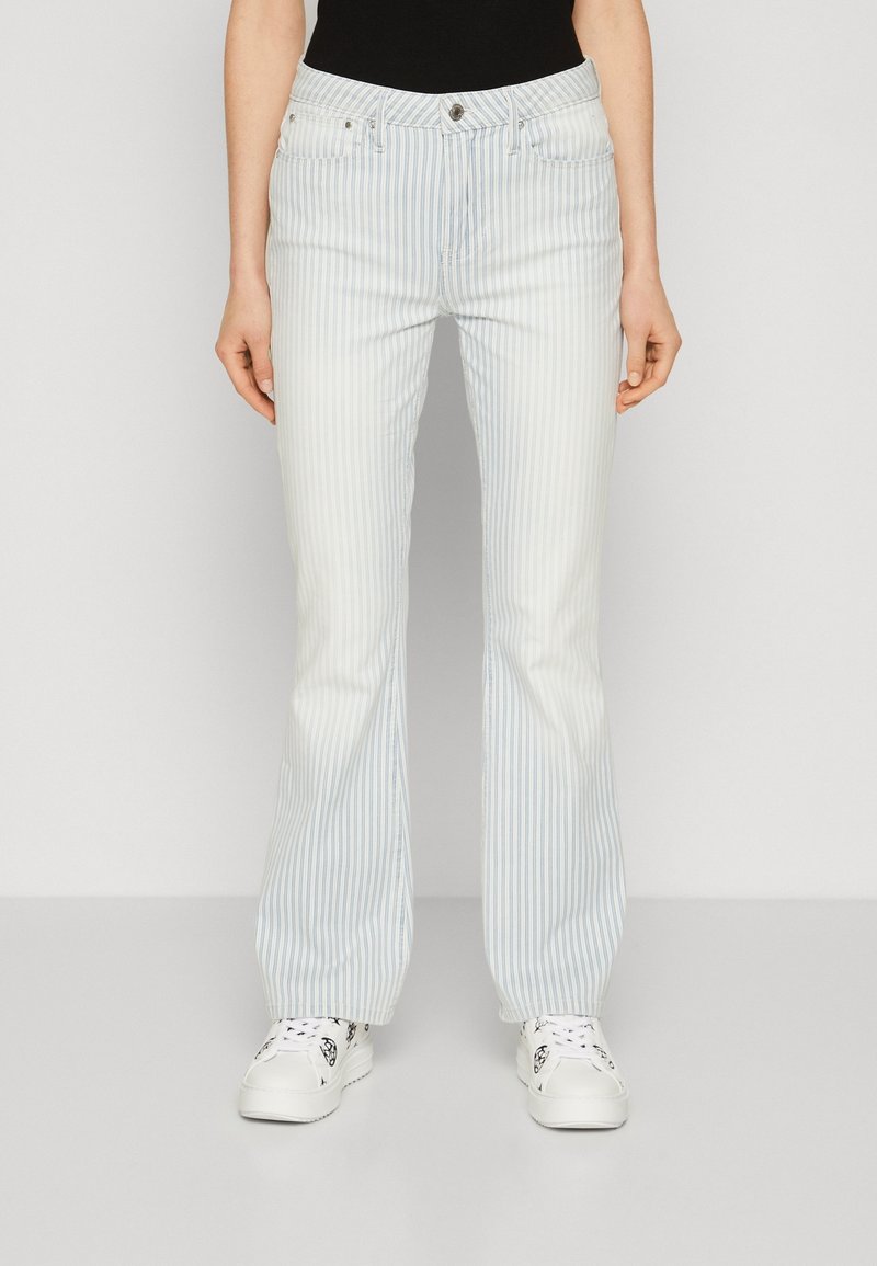 Personne portant un jean évasé à rayures verticales bleu clair et blanc ainsi que des baskets blanches sur un fond uni.