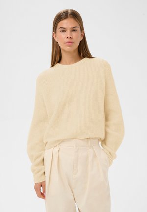 Vrouw met stijlbruin haar, gekleed in een lichtbeige gebreide trui en een hoge taille crème plissé broek, staand met één hand in de zak.