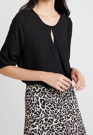 Woman wearing a black blouse with a front slit neckline and a high-waisted leopard print skirt, hands adjusting clothing.