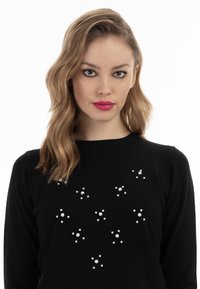 Schwarzer Pullover mit rundem Ausschnitt und verstreuten weißen Perlenverzierungen in verschiedenen Größen auf der Vorderseite. Weicher Strickstoff.