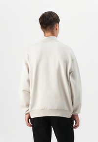 Švarkas su ilgomis rankovėmis šviesiai beige spalvos, turintis laisvą siluetą, išraižytas užbaigimas prie rankovių ir švelni tekstūra. Nėra matomų logotipų ar raštų.