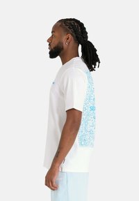 T-shirt blanc à manches courtes avec un motif paisley bleu sur le côté gauche et du texte au-dessus. Fabriqué en coton doux.