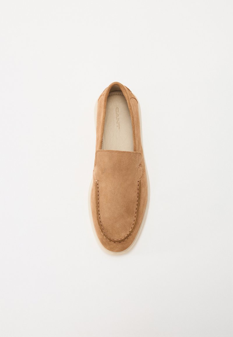 Chaussure slip-on en suede beige avec un bout arrondi, présentant des accents cousus et une semelle lisse de couleur claire. Design minimaliste sans quincaillerie visible.