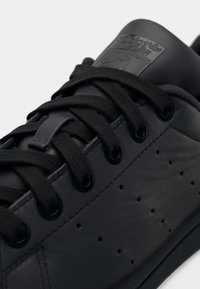 Černá kožená teniska s černými tkaničkami, perforovaným bočním detailem a značkovou štítkovou nálepkou na jazyku na bílém pozadí.