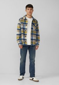 Koszula flanelowa w niebieską, żółtą i beżową kratkę, z kołnierzykiem i kieszonką na piersi; zestawiona z dżinsami i białymi trampkami.