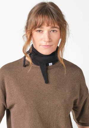 Ung kvinde med lyst brunt hår iført en brun top og en sort strikket halsvarmer mod en ensfarvet hvid baggrund.