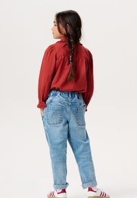 Czerwona, fakturowana koszula z długim rękawem i marszczonym karczkiem, w połączeniu z jasnoniebieskimi spodniami z wysokim stanem, które mają dwie tylne kieszenie i podwinięte mankiety.