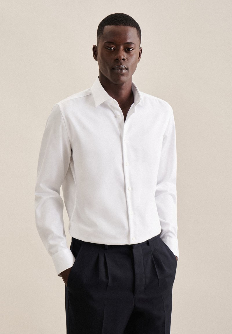 Chemise blanche à manches longues avec un col boutonné, en tissu doux et à coupe ajustée. Associée à un pantalon plissé foncé.