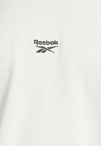 Bílá tkanina s jemnou texturou, v levém horním rohu s černě vyšívaným logem Reebok, prezentující elegantní design.