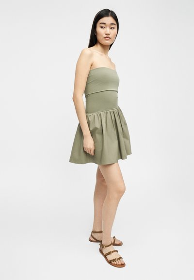 Νεαρή γυναίκα με μακριά σκούρα μαλλιά, που φοράει φόρεμα χωρίς τιράντες σε ελιά πράσινο και καφέ επίπεδα σανδάλια, στέκεται μπροστά σε απλό φωτεινό φόντο.