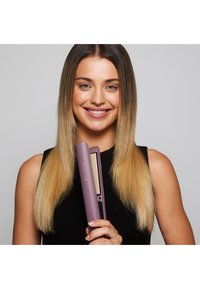 Lilla hårstrykare med guldplattor, hålls av en person med långt, rakt, ljusbrunt hår. Slät yta och elegant design.