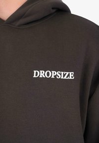 Sudadera marrón oscuro hecha de un tejido suave, con un logo blanco "DROPSIZE" en el pecho, de diseño clásico y con una capucha amplia.