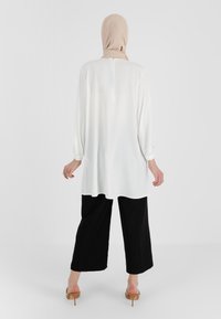 Weiße Tunika mit langen Ärmeln, lockerem Schnitt, plissierten Ärmeln und Reißverschluss hinten. Kombiniert mit schwarzen, weit geschnittenen Hosen und beigen High Heels.