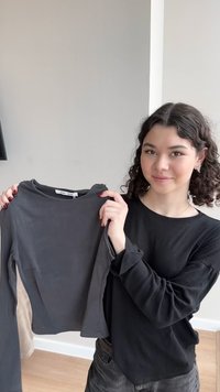 Νεαρή γυναίκα με σγουρά μαλλιά κρατάει ένα μακρυμάνικο πουκάμισο σκούρου γκρι χρώματος σε εσωτερικό χώρο, χαμογελώντας ελαφρώς προς την κάμερα.