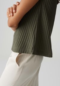 Top a coste verde oliva con spacchi laterali, abbinato a pantaloni color crema. Concentrati sulla texture e sulla vestibilità senza abbellimenti.