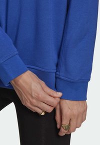 Felpa blu con polsini e orlo a coste. Primo piano di mani che aggiustano la manica, con anelli d'oro. Texture morbida, design casual.