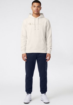 Giovane uomo in piedi che indossa una felpa con cappuccio beige di Champion, pantaloni della tuta blu navy di Champion e sneakers bianche di Champion su uno sfondo neutro.