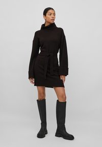 Vestido de malha preto com uma golinha alta, mangas longas e uma cintura atada. Apresenta um acabamento texturizado e uma barra ligeiramente acima do joelho.