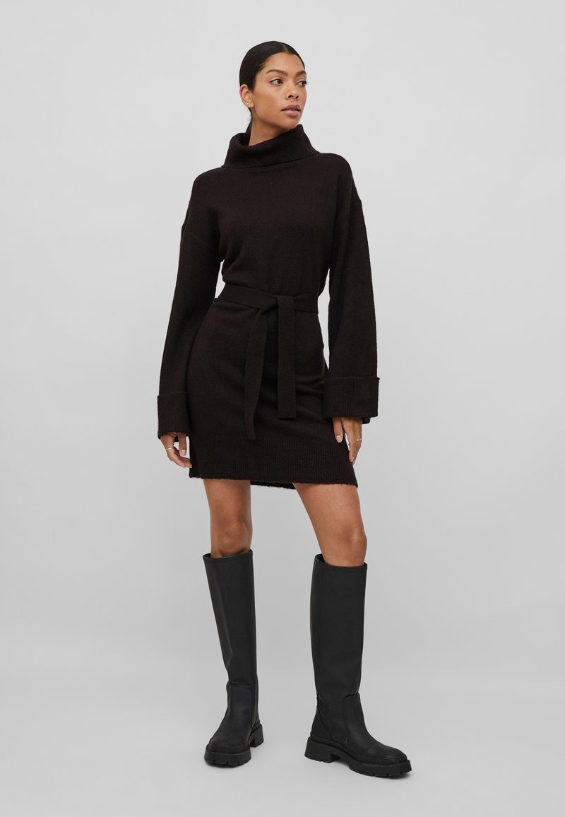 Vestido de malha preto com uma golinha alta, mangas longas e uma cintura atada. Apresenta um acabamento texturizado e uma barra ligeiramente acima do joelho.