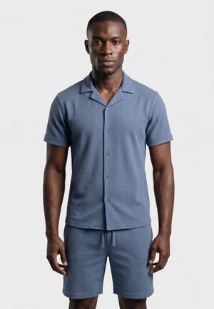 Homme debout, face à nous, portant une chemise bleue à manches courtes avec des boutons et un short assorti sur un fond gris uni.