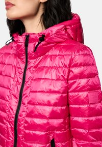 Woman wearing a bright pink quilted jacket with a hood and black zipper details, shown from the shoulder up, looking left.