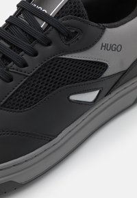 Baskets de sport noires avec un upper en mesh, des accents synthétiques et des détails gris. Présente une semelle texturée et des lacets plats. Logo sur le côté.