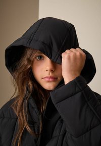 Czarna puchowa kurtka z kapturem i pikowaną teksturą. Modelka częściowo zakryta, eksponująca zamek błyskawiczny i przeszycia.