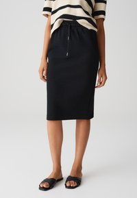 Schwarzer Strickrock mit gerader Silhouette, elastischem Bund und Kordelzugdetail. Kombiniert mit schwarzen Riemchenslippers.