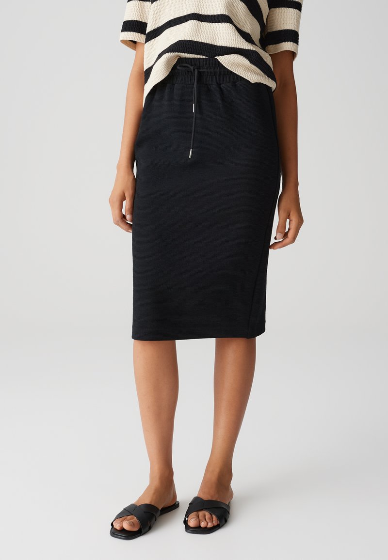 Schwarzer Strickrock mit gerader Silhouette, elastischem Bund und Kordelzugdetail. Kombiniert mit schwarzen Riemchenslippers.