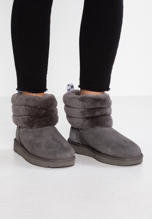 Personne portant des bottines grises doublées en peluche avec des poignets rabattus et un pantalon noir ajusté, debout sur un sol blanc.