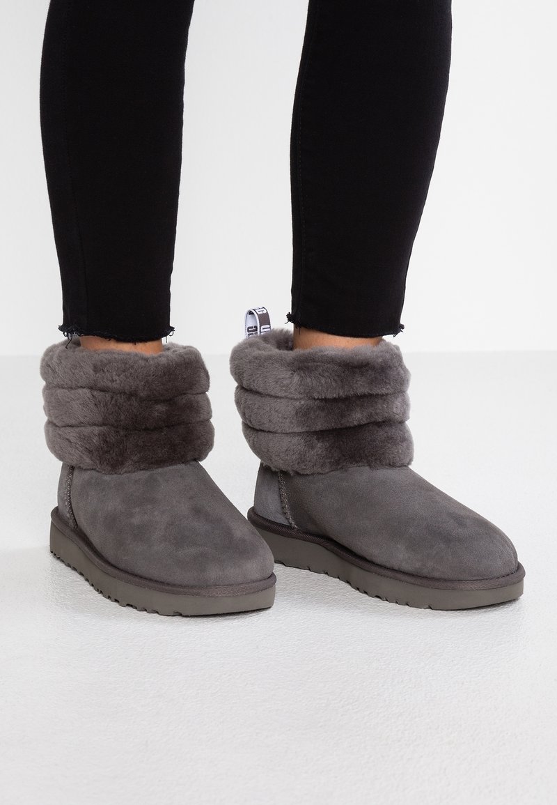 Personne portant des bottines grises doublées en peluche avec des poignets rabattus et un pantalon noir ajusté, debout sur un sol blanc.