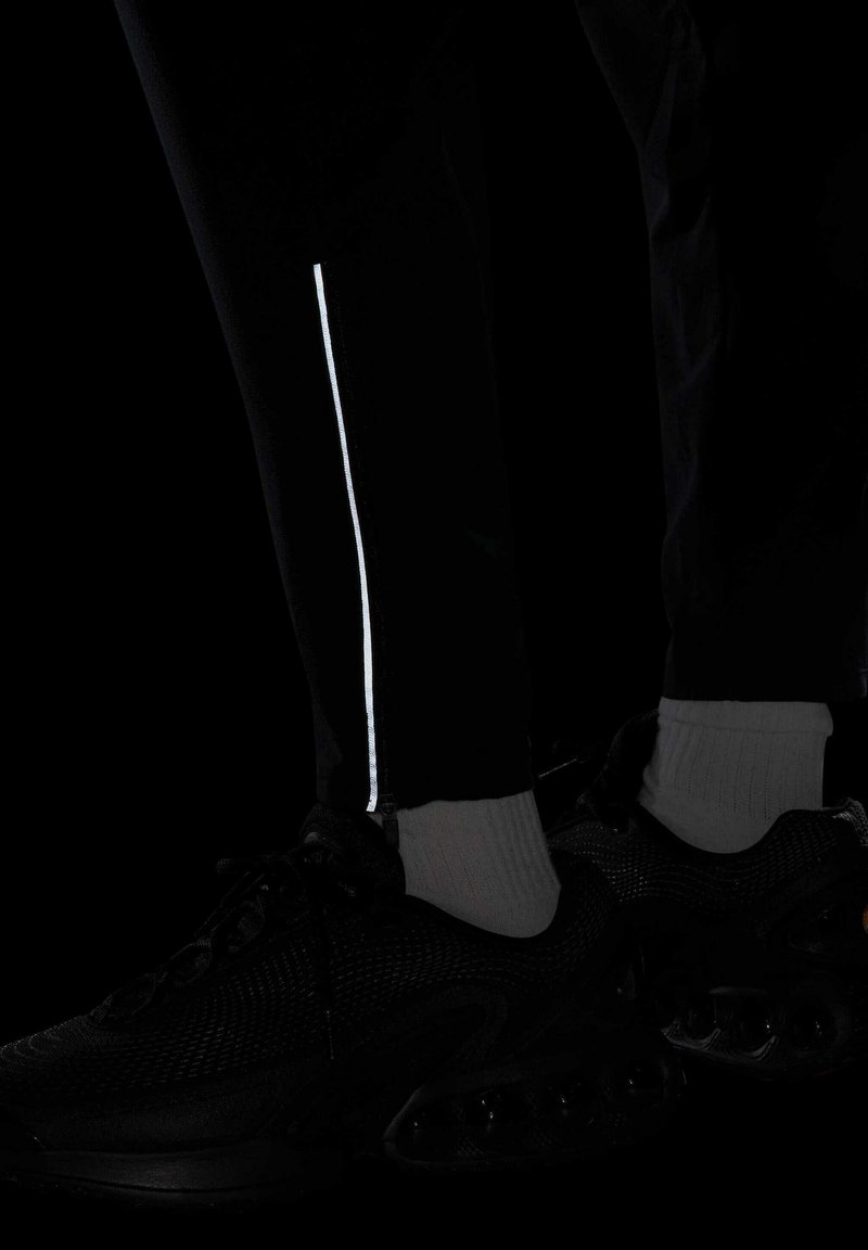 Pantalons de sport noirs avec une bande réfléchissante blanche sur le côté, associés à des baskets noires et des chaussettes mi-mollet grises. La texture semble lisse.