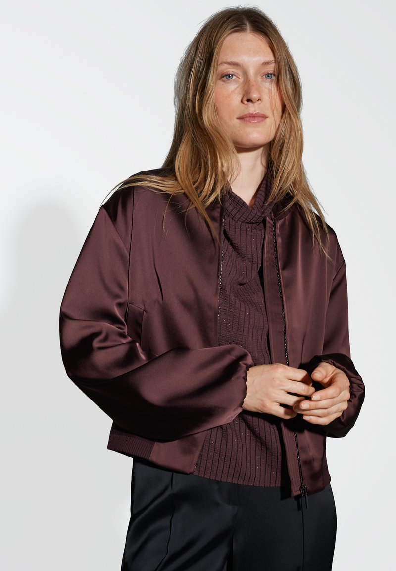 Giacca bomber in satin bordeaux con zip frontale, polsini a coste e vestibilità rilassata, indossata sopra un top in maglia testurizzata.
