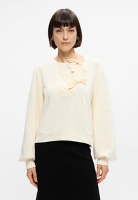Sweat-shirt couleur crème avec de longues manches bouffantes, ornée de deux nœuds pêche clair au niveau du col et d'un design en tissu texturé.