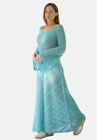 Długa, jasnoniebieska dzianinowa sukienka z ukośnym wzorem, z długimi rękawami i luźnym krojem, noszona na białym obuwiu.
