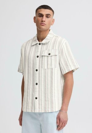 Mann trägt ein kurzärmeliges, beige-weiß gestreiftes Hemd mit Knopfleiste und Brusttasche sowie hellblaue Jeans vor einfachem Hintergrund.