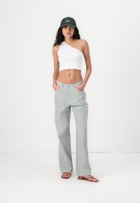 Witte one-shoulder crop top, groen gestreepte wijde broek en bruine sandaal. Model draagt een donkergroene pet. Eenvoudig, aansluitend ontwerp.