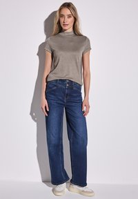 Gray short-sleeve top with a high neckline paired with wide-leg dark blue jeans. Model stands against a light background, wearing white sneakers.