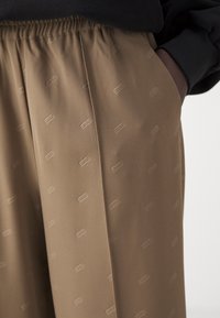 Beige plisserte bukser med elastisk liv, med et subtilt preget logomønster. Stoffet virker glatt og lett.