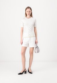 Top bianco a maniche corte con un motivo testurizzato, abbinato a pantaloni corti bianchi e sandali con tacco a blocco in pelle verniciata nera. Tiene una borsa argento.