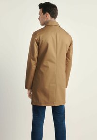 Brązowy, do kolan płaszcz o prostym kroju, z długimi rękawami i małym kołnierzykiem. Gładka tekstura, noszony na niebieskie dżinsy.