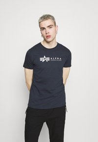 Marineblå bomulls t-skjorte med en hvit logo "ALPHA" med stiliserte bokstaver og striper over brystet. Rund hals, korte ermer.