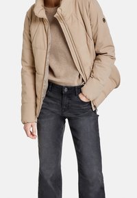 Beige pufferväst med synliga sömmar, dragkedja framtill och rund logopatch. Kombinerad med en strukturerad beige stickad tröja och mörka jeans.