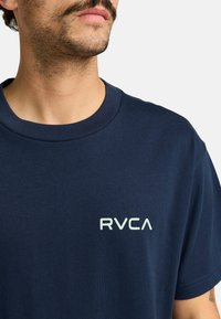 Marinblå bomullströja med rund halsringning, korta ärmar och en liten RVCA-logotyp i ljusgrön på vänster bröst. Slät textur.