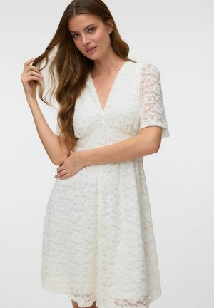 Mujer con cabello largo y castaño que lleva un vestido blanco de encaje con mangas cortas y largo hasta la rodilla, posando con una mano en el cabello y la otra en la cintura.
