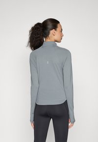 Grå langermet treningsgenser med høy krage. Har en sømløs design, glatt tekstur og en subtil logo på baksiden. Svart leggings er synlig.